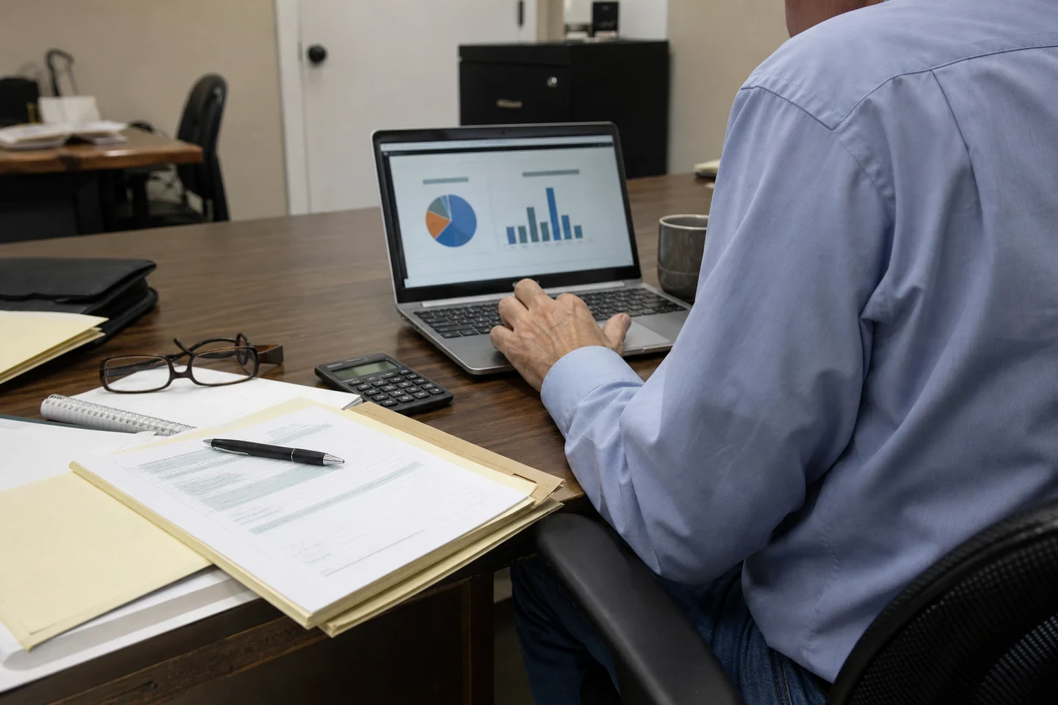 Man in a light blue shirt at an office desk reviewing pie and bar charts on a laptop, with folders, paperwork, a pen, a calculator and reading glasses beside him